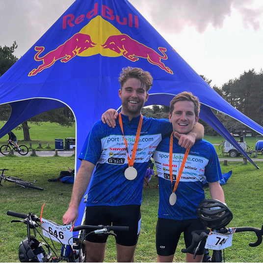 Two cyclists with medals standing in front of a Red Bull tent at a cycling event